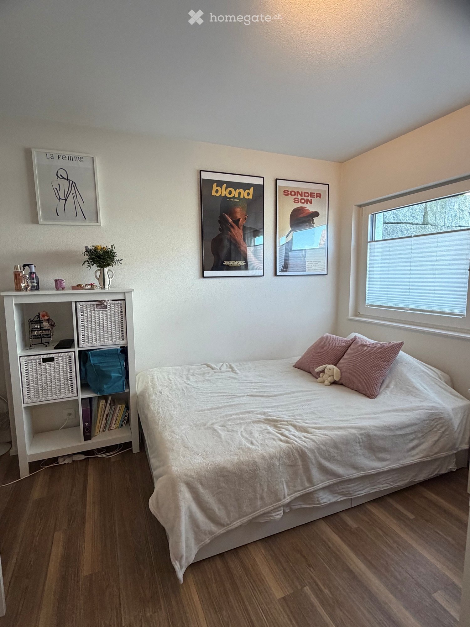 Cozy bedroom with a white bed, pink pillows, a small shelf with books and decorative items, and framed posters on the wall.