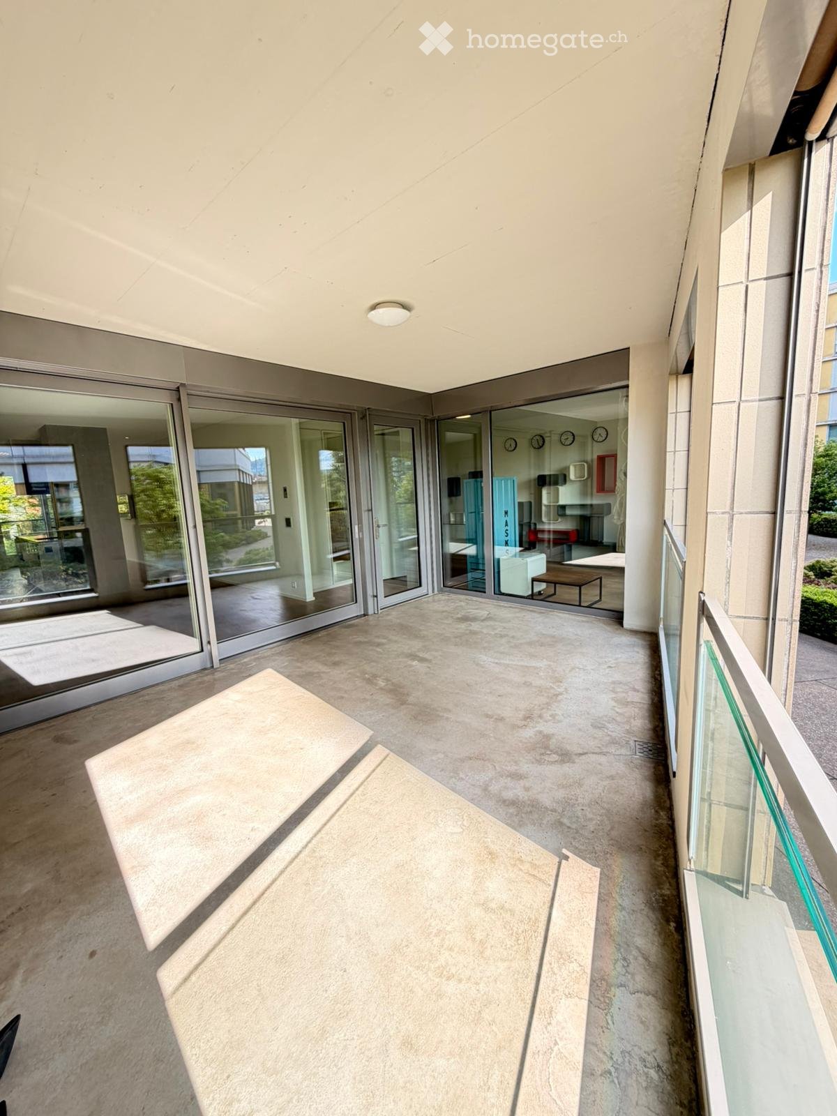Modern balcony with concrete floor, glass sliding doors, and a glass railing, leading to an indoor area with visible furniture.