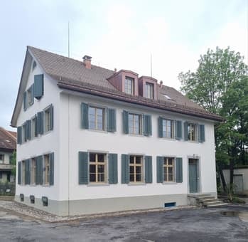 House. Two-story house with white walls, green shutters, and a red roof. The house features multiple windows and a small staircase leading to the entrance.