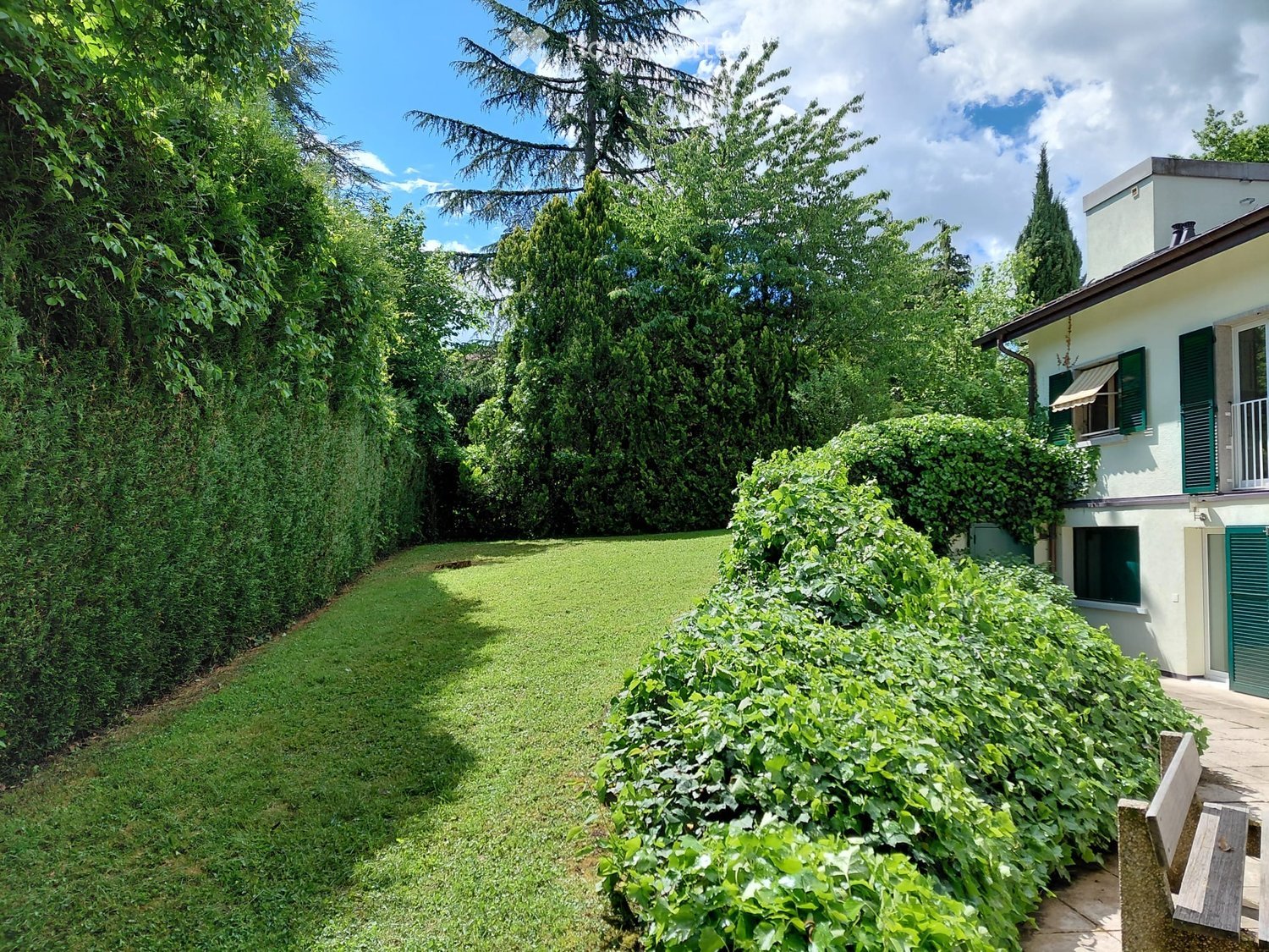 Well-maintained garden with a trimmed hedge, a neatly mowed lawn, and a pathway leading to a house with green shutters and a small balcony.