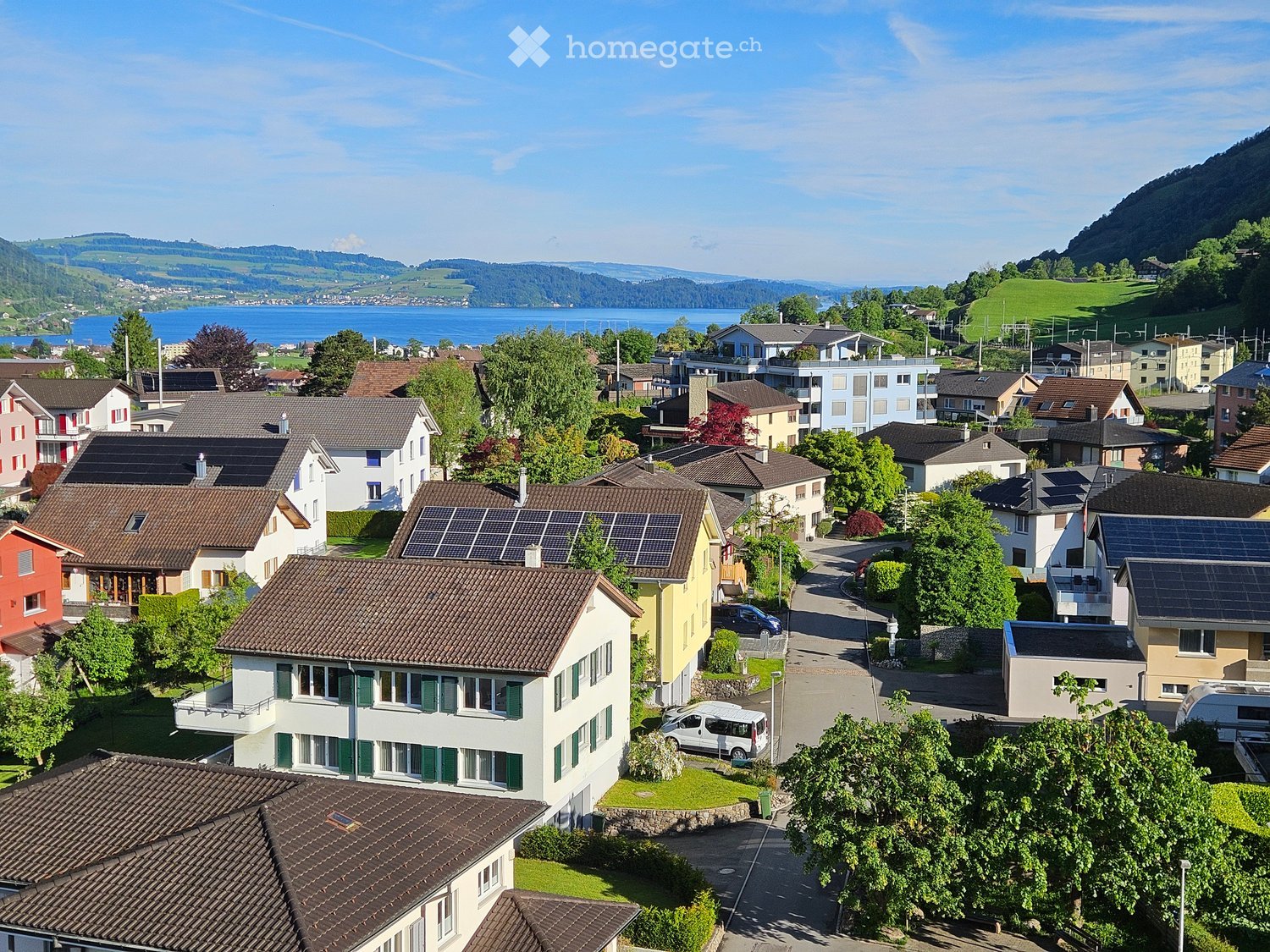Aerial view of a residential neighborhood with multiple houses, solar panels on some roofs, and a scenic lake and mountains in the background.
