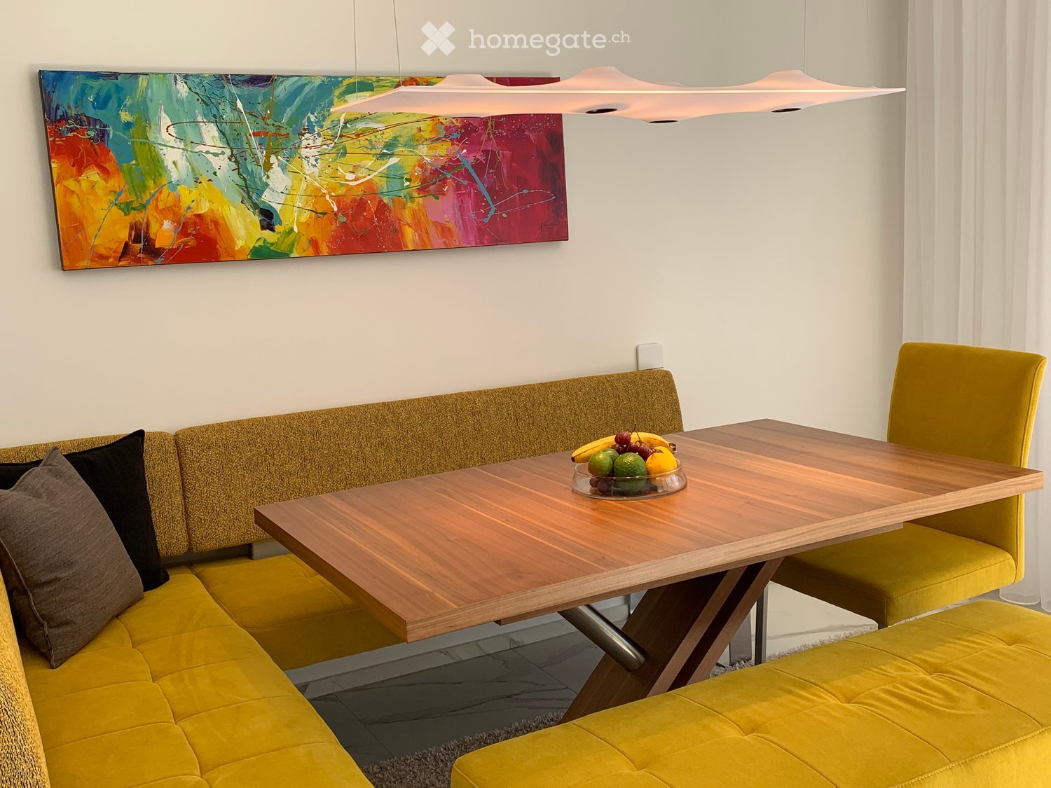 Furnished Room. Bright living room with a yellow sofa, wooden table, and modern pendant light. A colorful abstract painting hangs on the wall, and a fruit bowl sits on the table.