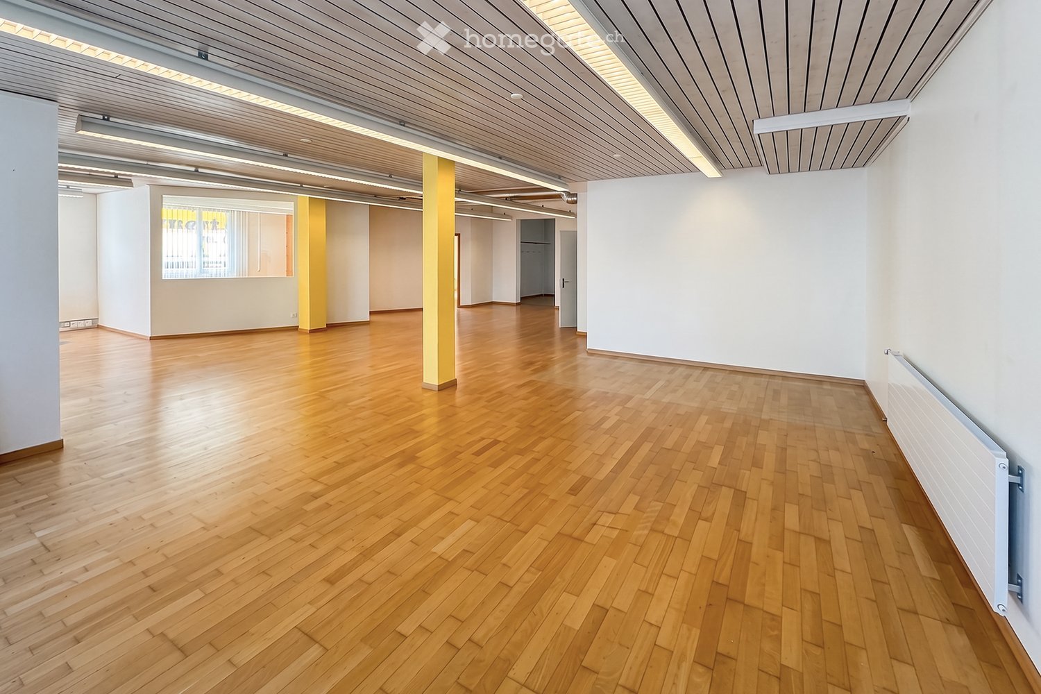Unfurnished Room. Empty room with wooden flooring, white walls, and a central column. The space is well-lit with natural light from a window and recessed ceiling lighting.