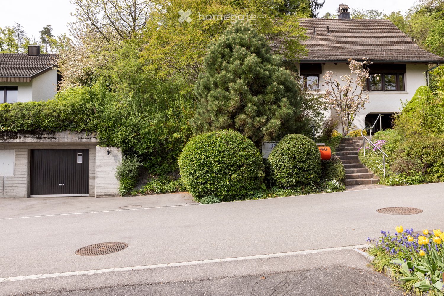 Two-story house with a tiled roof, white exterior, and a black garage door. The house is surrounded by lush greenery and has a staircase leading up to the entrance.