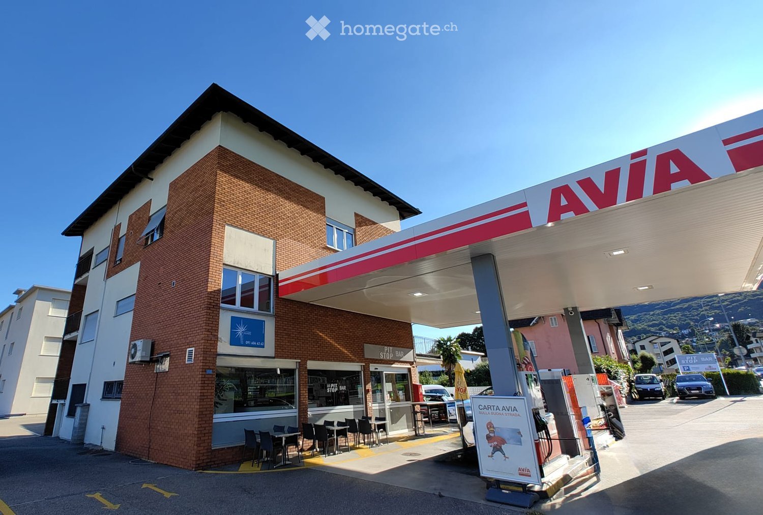 Building. A two-story commercial building with a brick facade and a large 'AVIA' sign, featuring a gas station canopy and a small seating area with tables and chairs.