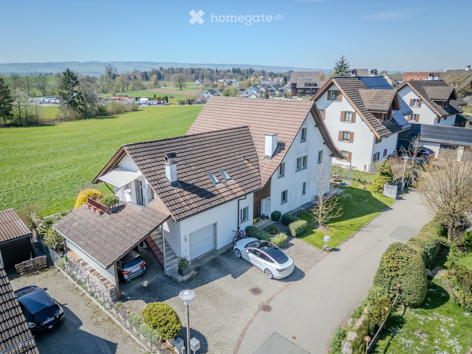 House. A two-story house with a tiled roof, white exterior, and a garage. The property includes a driveway with parked cars and a well-maintained garden.