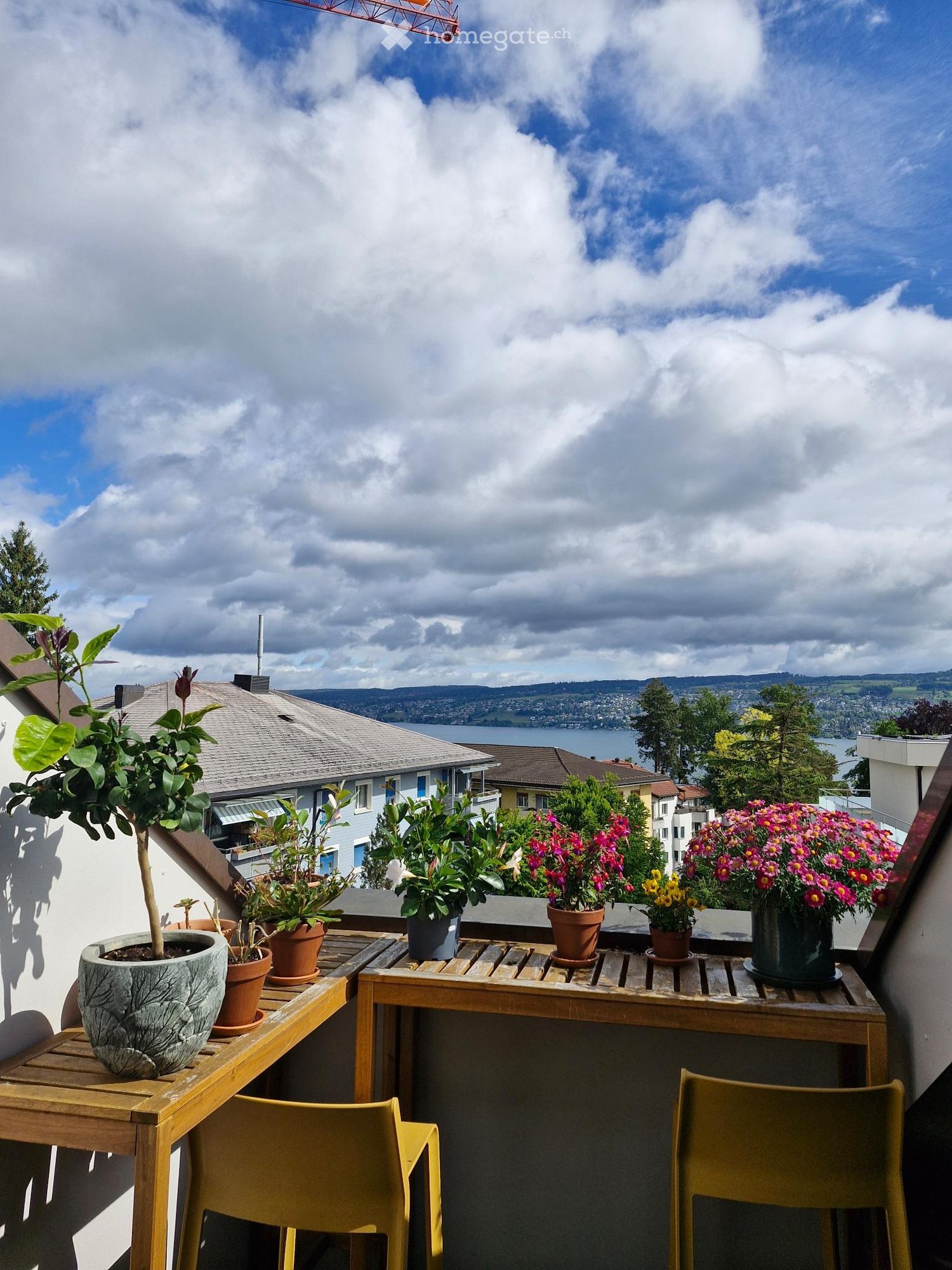 Balcony / Terrace. Balcony with wooden table, yellow chairs, and several potted plants, overlooking a scenic view of a lake and distant hills.