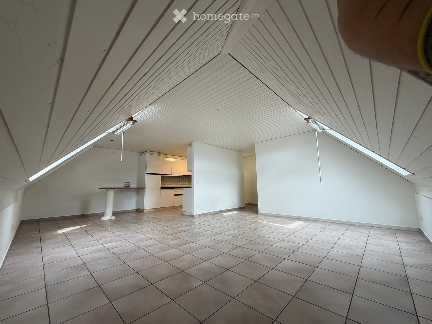 Bright, open attic space with slanted wooden ceilings, tiled floor, and a small kitchen area featuring cabinets, a refrigerator, and a table.