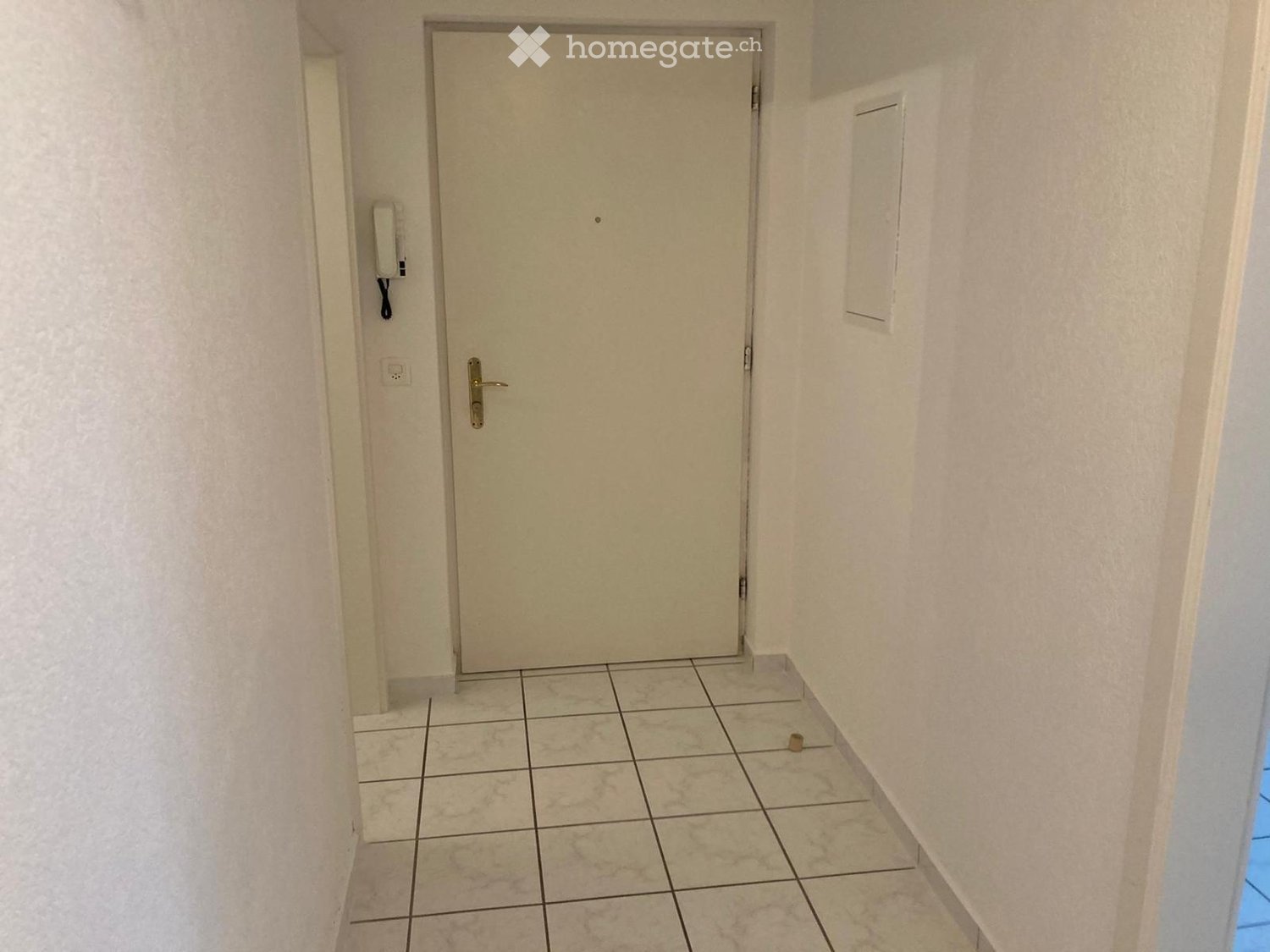 Corridor / Hallway. A narrow hallway with tiled flooring and light-colored walls, featuring a closed door at the end and a small intercom system mounted on the wall.
