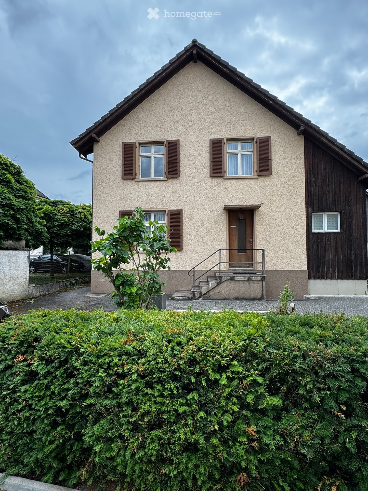 House. Two-story house with beige siding, wooden shutters, and a small staircase leading to the front door. The house is surrounded by greenery and has a well-maintained garden in front.