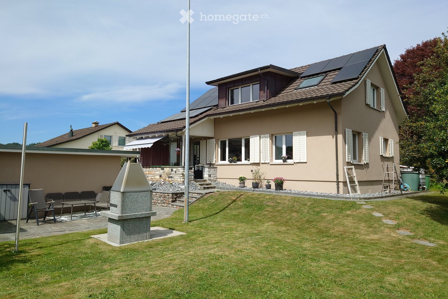 Garden / Outdoor Area. A well-maintained garden with a lawn, patio seating area, and a stone fire pit. The house features solar panels on the roof and multiple windows with shutters.