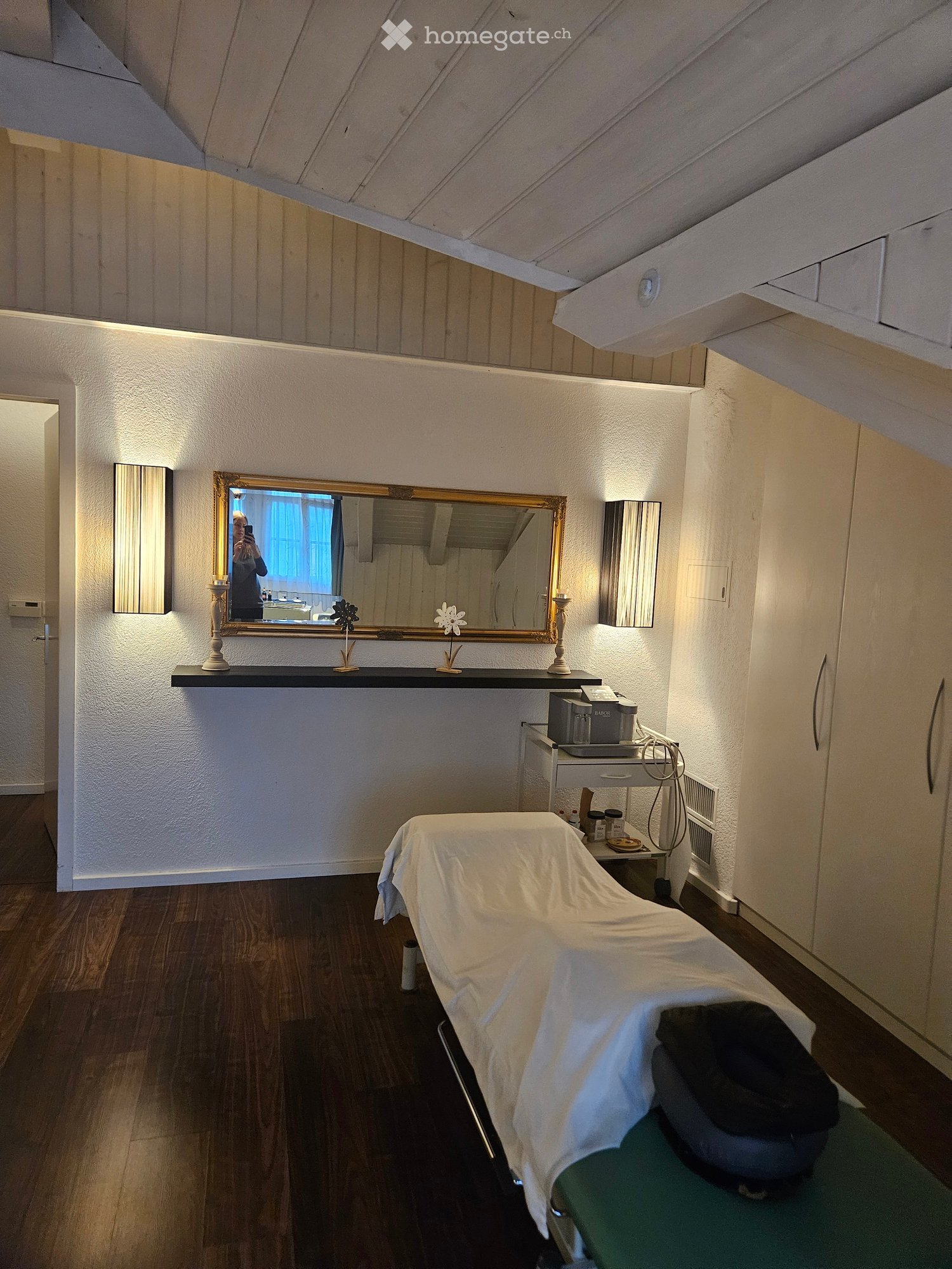 Furnished Room. A small, well-lit room with a treatment table, a mirror, a small table with items, and a cabinet.