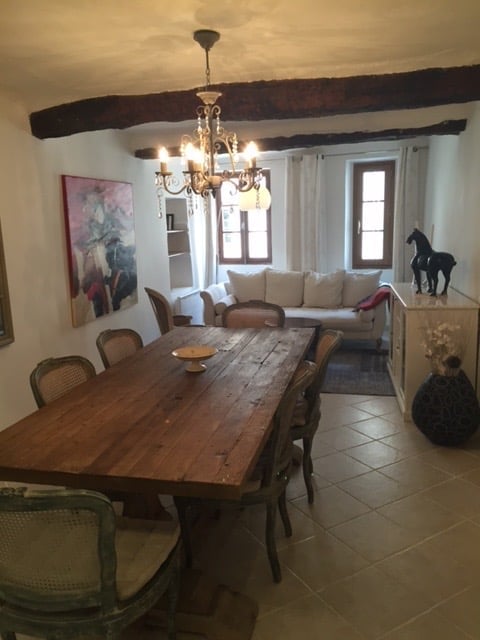 Furnished Room. A cozy, rustic living and dining area with exposed wooden beams, a chandelier, a sofa, a wooden dining table with chairs, a painting, a vase, and a decorative sculpture.