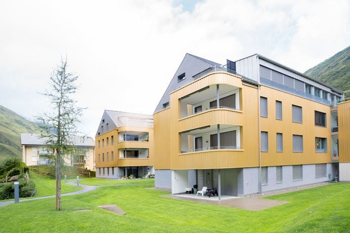 Building. Modern multi-story building with a combination of wood and concrete facade, featuring balconies and large windows. The building is surrounded by a well-maintained lawn and pathways.