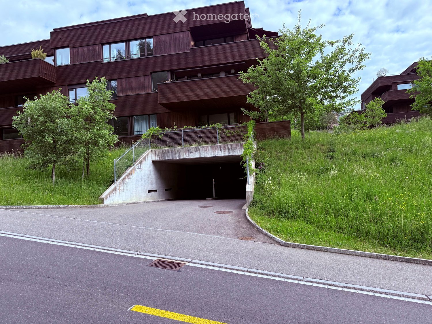 Building. Modern multi-story building with dark wooden facade, large windows, and balconies. A concrete ramp leads to an underground parking area surrounded by greenery.