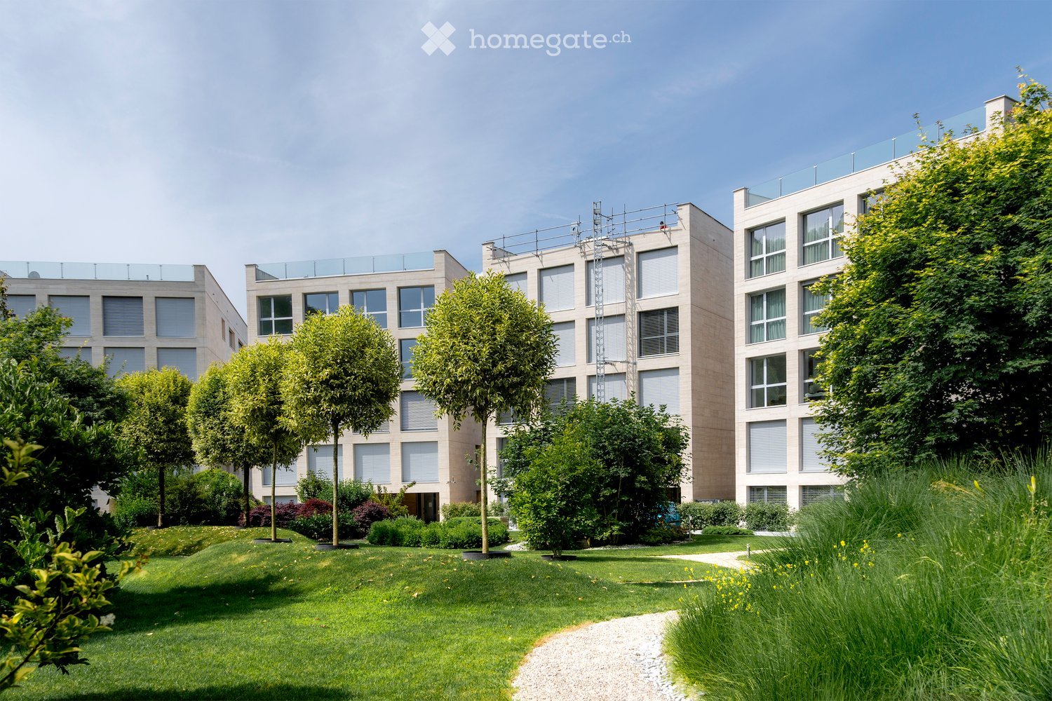 Building. Modern multi-story residential building with large windows, surrounded by a well-maintained garden with trees, shrubs, and a gravel pathway.