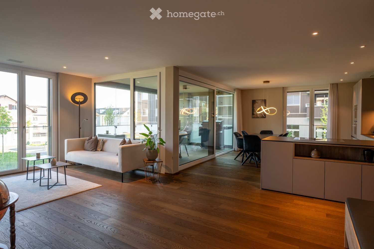 Furnished Room. Modern living and dining area with a white sofa, coffee table, side tables, dining table, chairs, and a kitchen island. Large windows and sliding glass doors lead to a balcony.