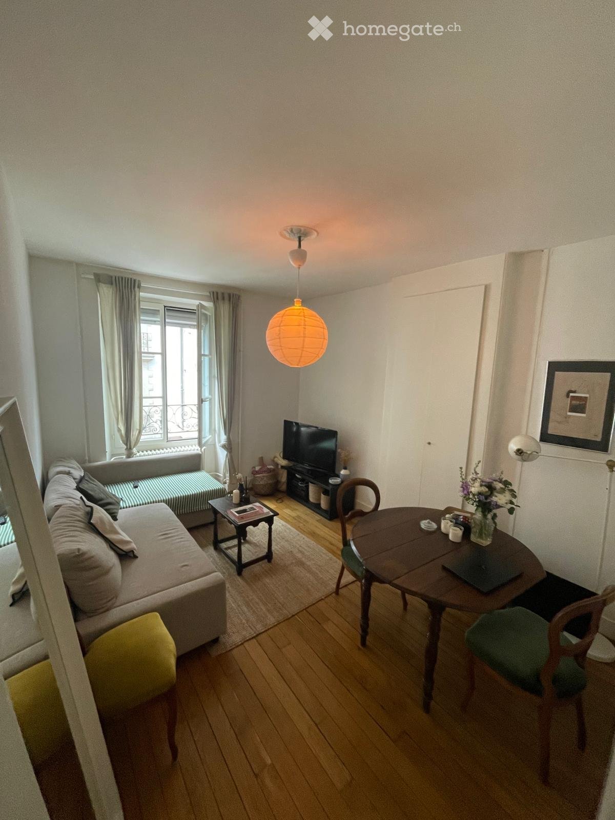 Furnished Room. A cozy living room with a sofa, small table, dining table, chairs, television, and a hanging paper lantern light fixture.
