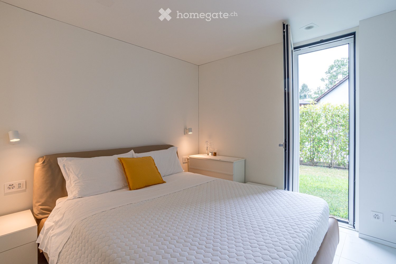Bedroom. Modern bedroom with a large bed, white bedding, a yellow throw pillow, and a nightstand with a lamp. Large sliding glass door leads to a green outdoor area.