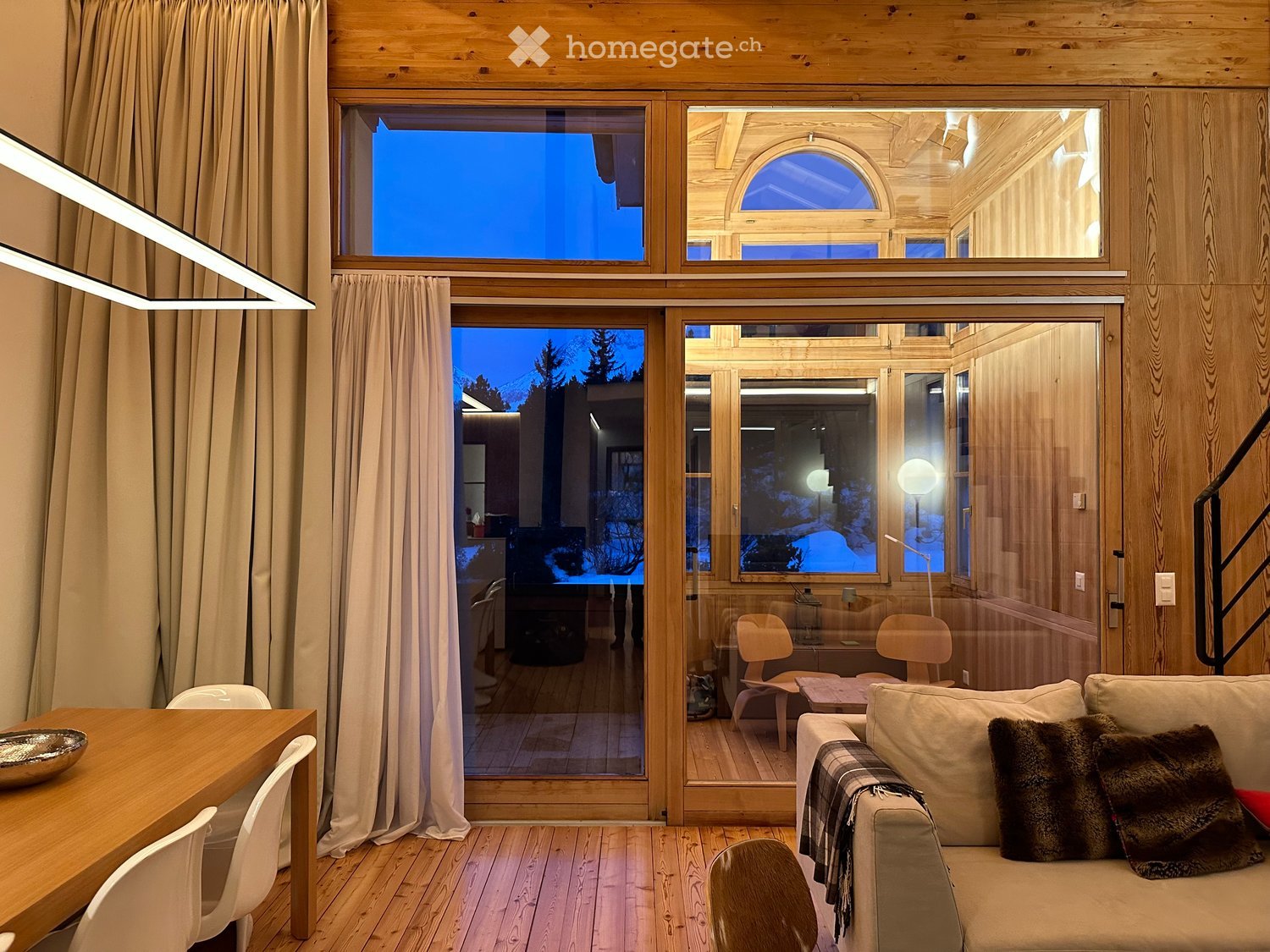 Furnished Room. Living room with wooden walls and floors, large windows, a sofa with pillows, a dining table with chairs, and a modern lamp.