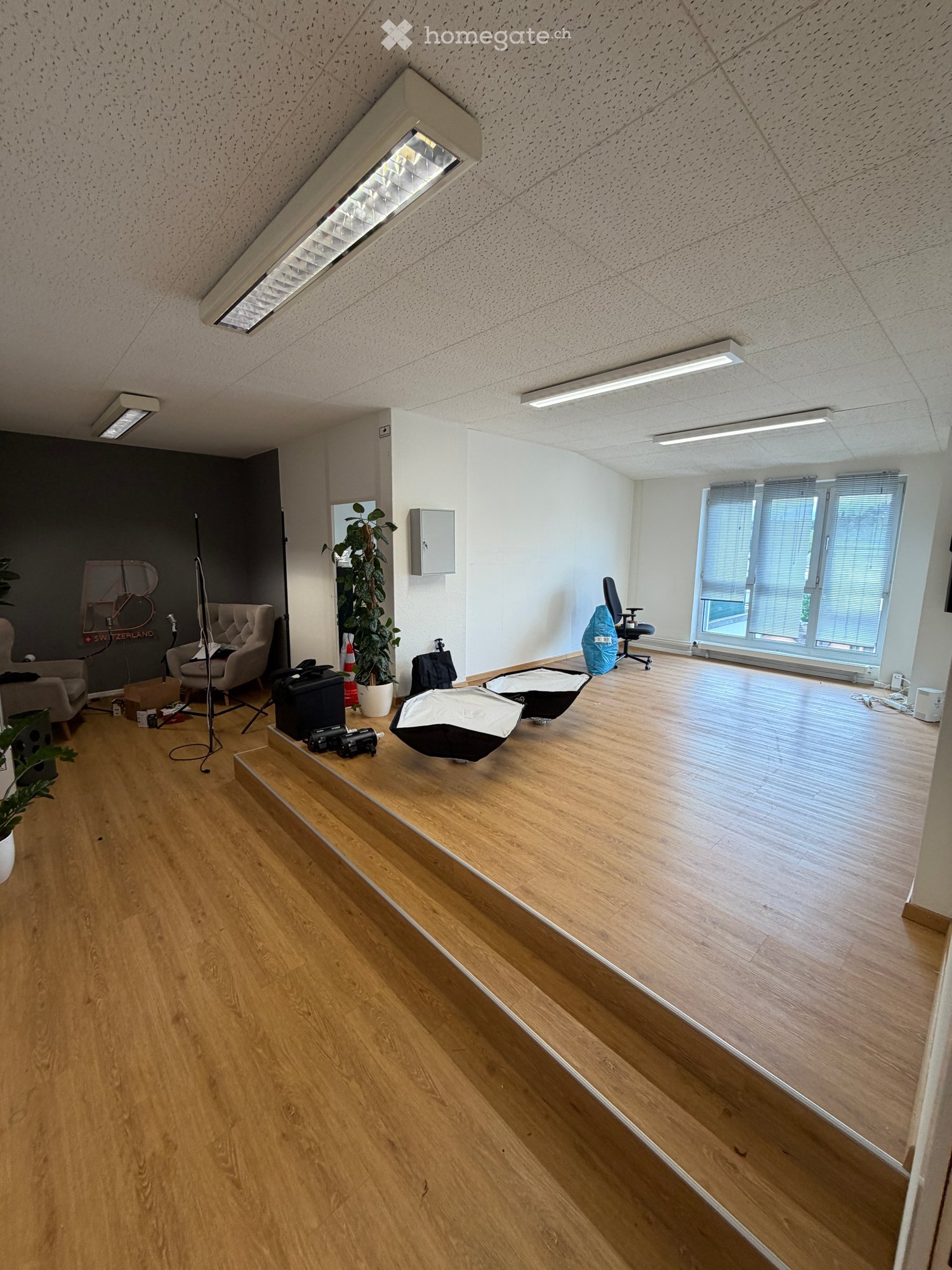 Furnished Room. Bright, open room with wooden flooring, large windows, and modern lighting. Contains a sofa, chair, office chair, plant, and various equipment including a camera and tripod.