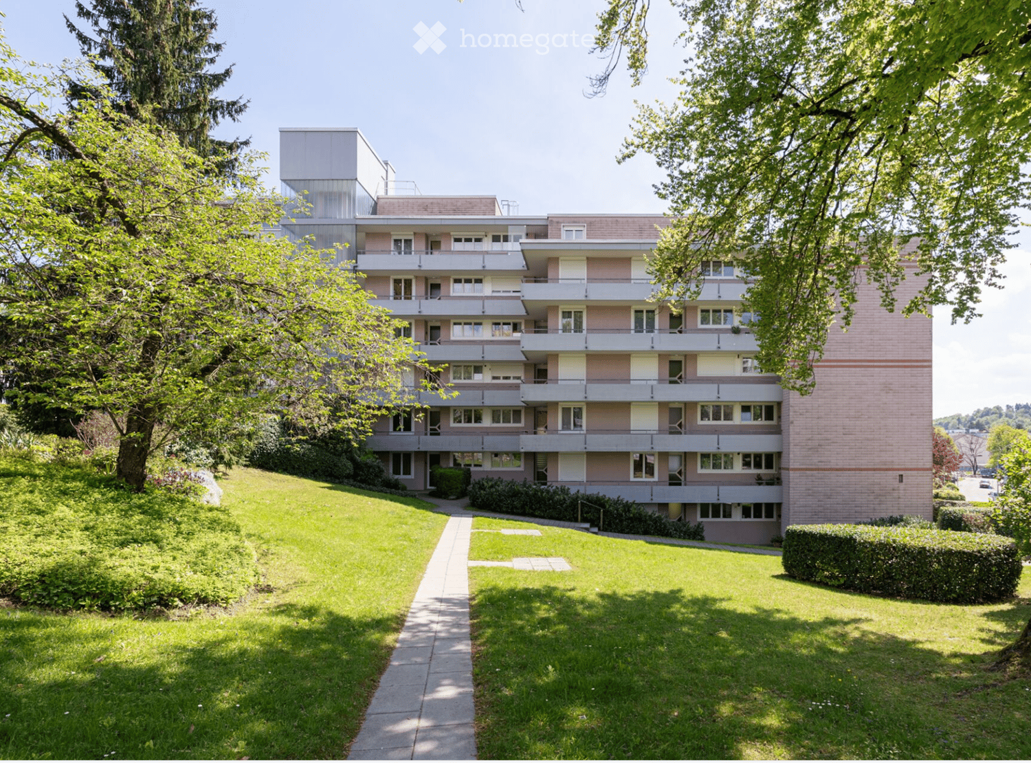 A multi-story apartment building with balconies, surrounded by greenery and a walkway leading to the entrance.