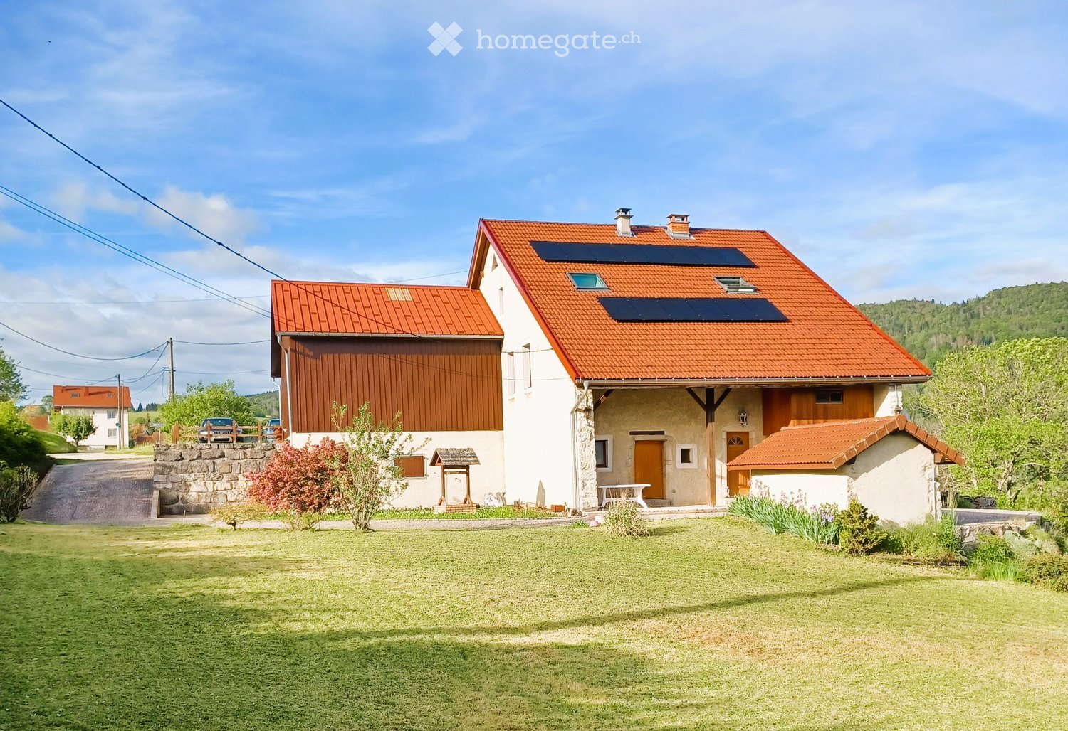 House. Two-story house with red-tiled roof, solar panels, stone and wood exterior, surrounded by a well-maintained lawn and garden area.