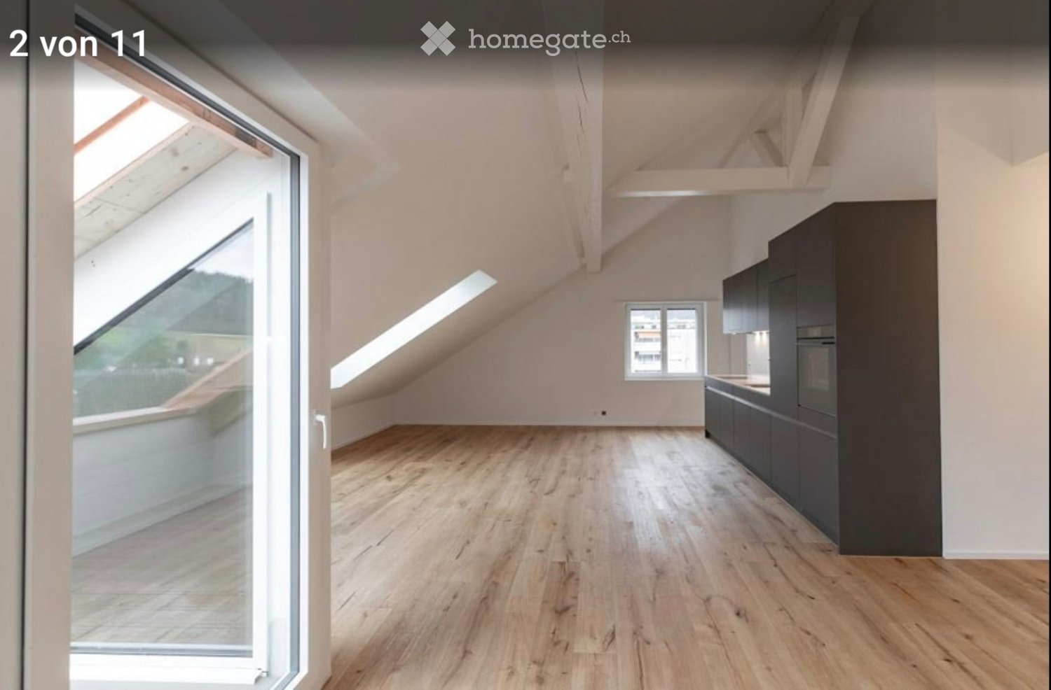 Unfurnished Room. Bright, open attic space with wooden flooring, white walls, and modern dark grey kitchen cabinets. Large skylight and window provide natural light.