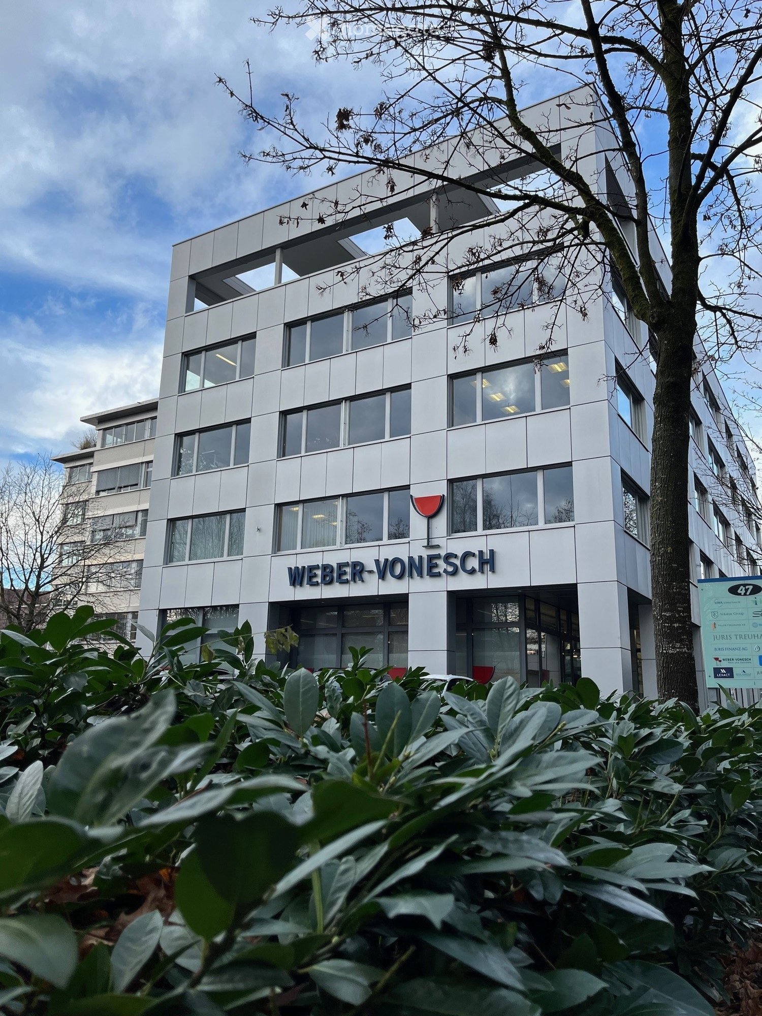Building. Modern multi-story building with large windows and a sign reading "WEBER-VONESCH" on the facade, surrounded by greenery and trees.