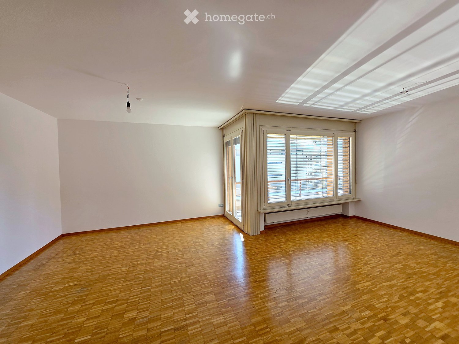 Empty room with wooden flooring, white walls, large windows with blinds, and a hanging light fixture.