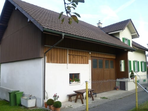House. Two-story house with a brown tiled roof, white and brown siding, and a wooden garage door. The front yard includes a small wooden picnic table, benches, and potted plants.