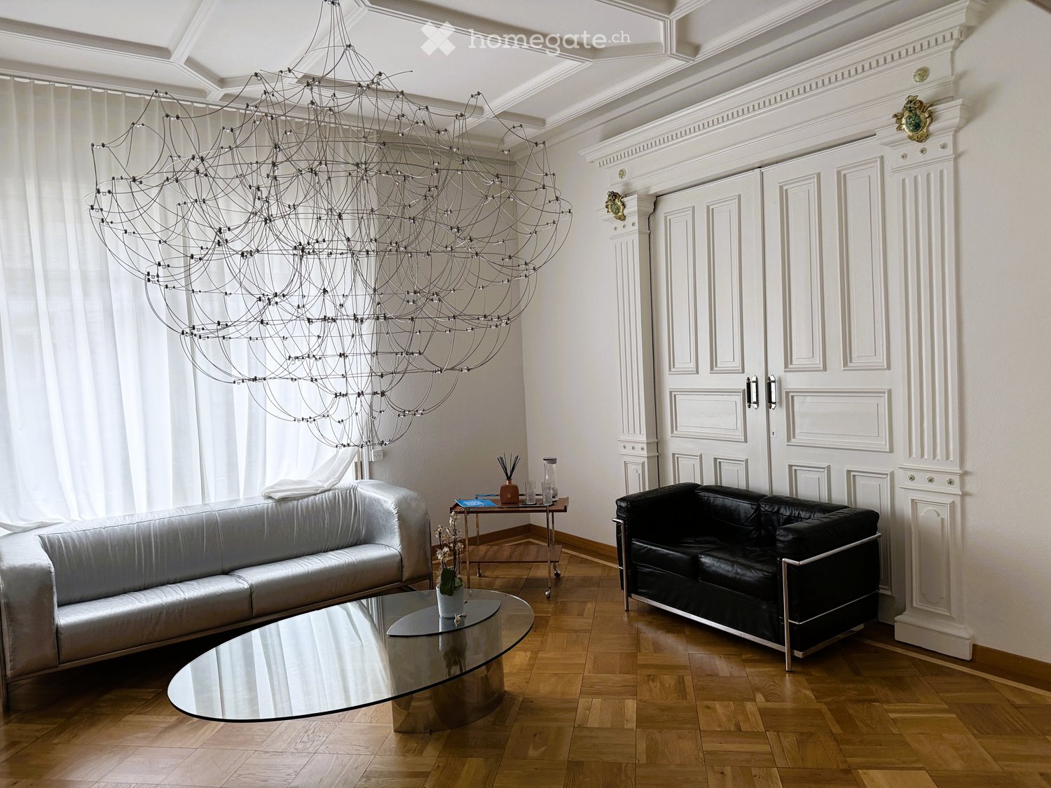 Living Room. Elegant living room with a large, modern chandelier, two sofas (one silver, one black), a glass coffee table, a small side table with a vase, and a large window with white curtains.