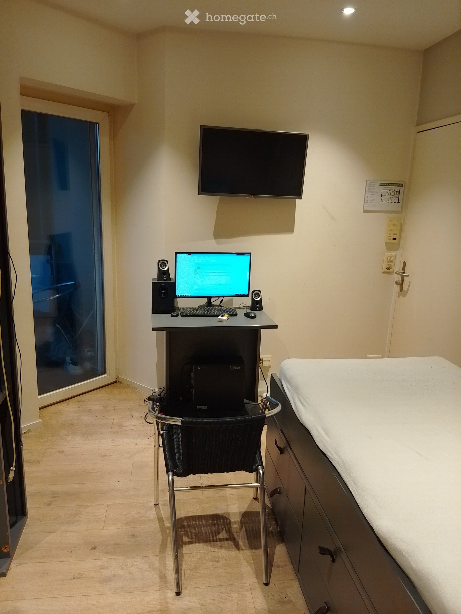 A room with a desk, computer, chair, bed, and a television mounted on the wall.