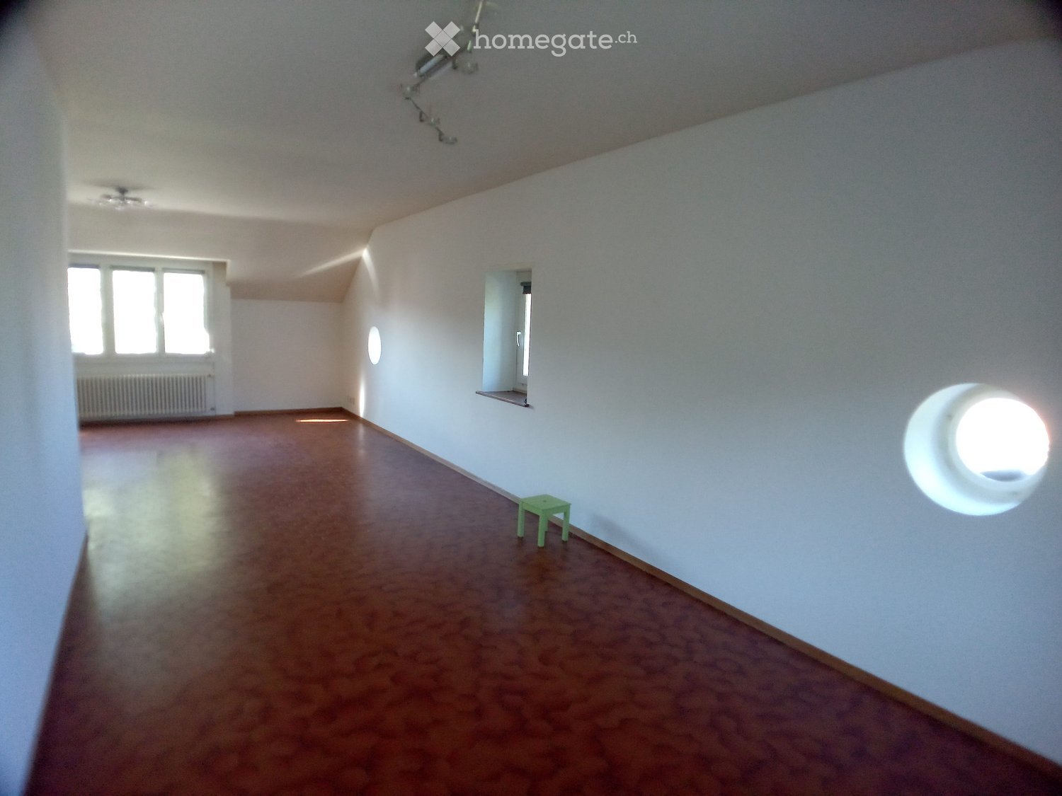 Empty room with wooden flooring, white walls, and large windows allowing natural light. A small green stool is placed near the wall.