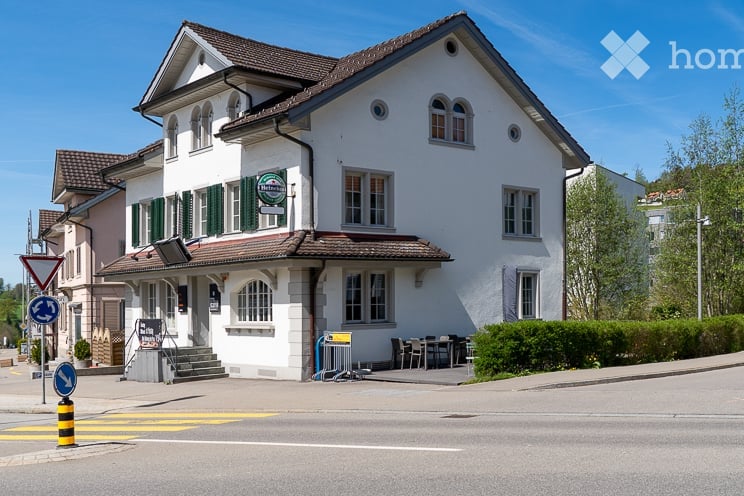 Building. A two-story white building with green shutters and a brown roof, featuring a small outdoor seating area with tables and chairs.