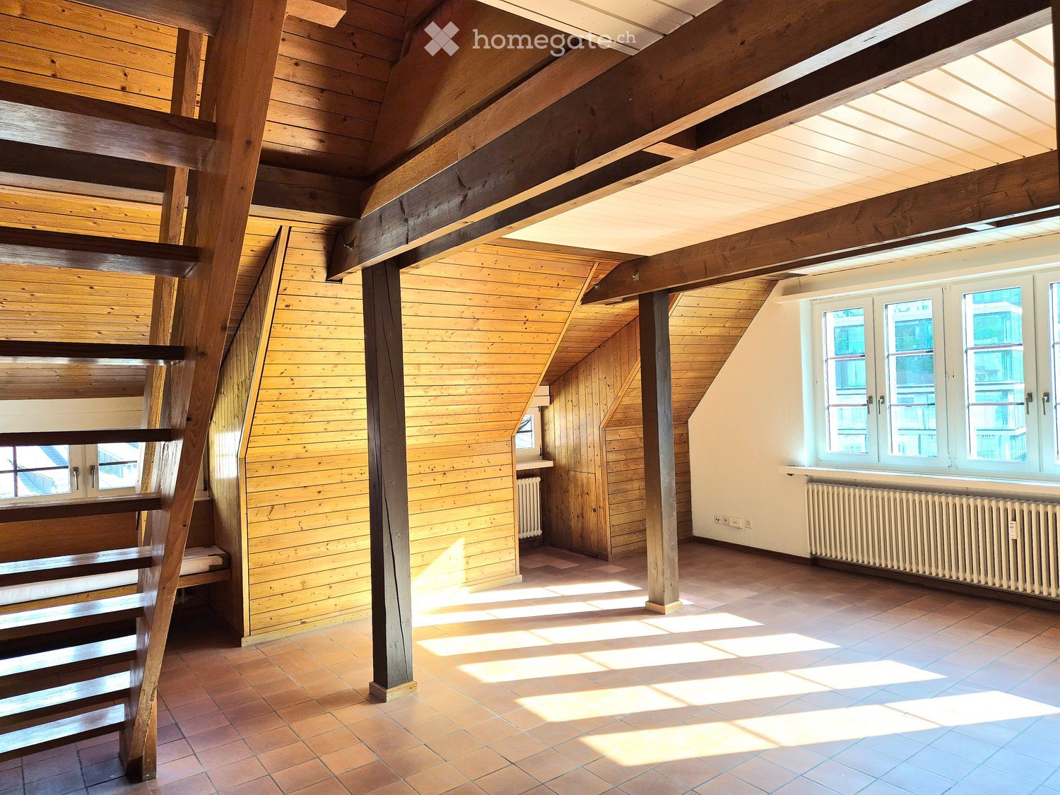 An unfurnished attic room with wooden walls, exposed wooden beams, a wooden staircase, and large windows allowing natural light to fill the space.
