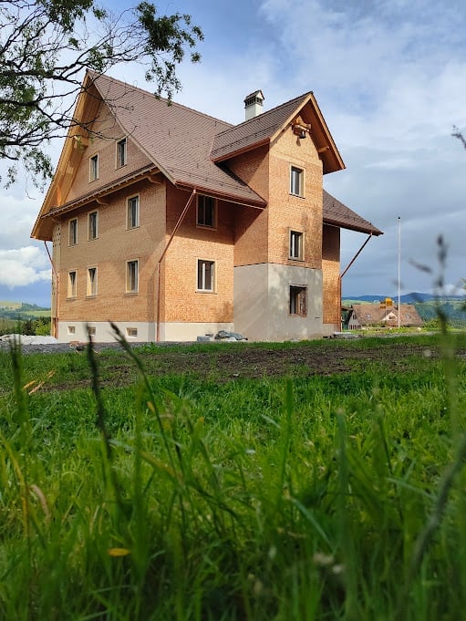 House. A two-story house with a brown roof and beige walls, surrounded by a grassy area and trees.