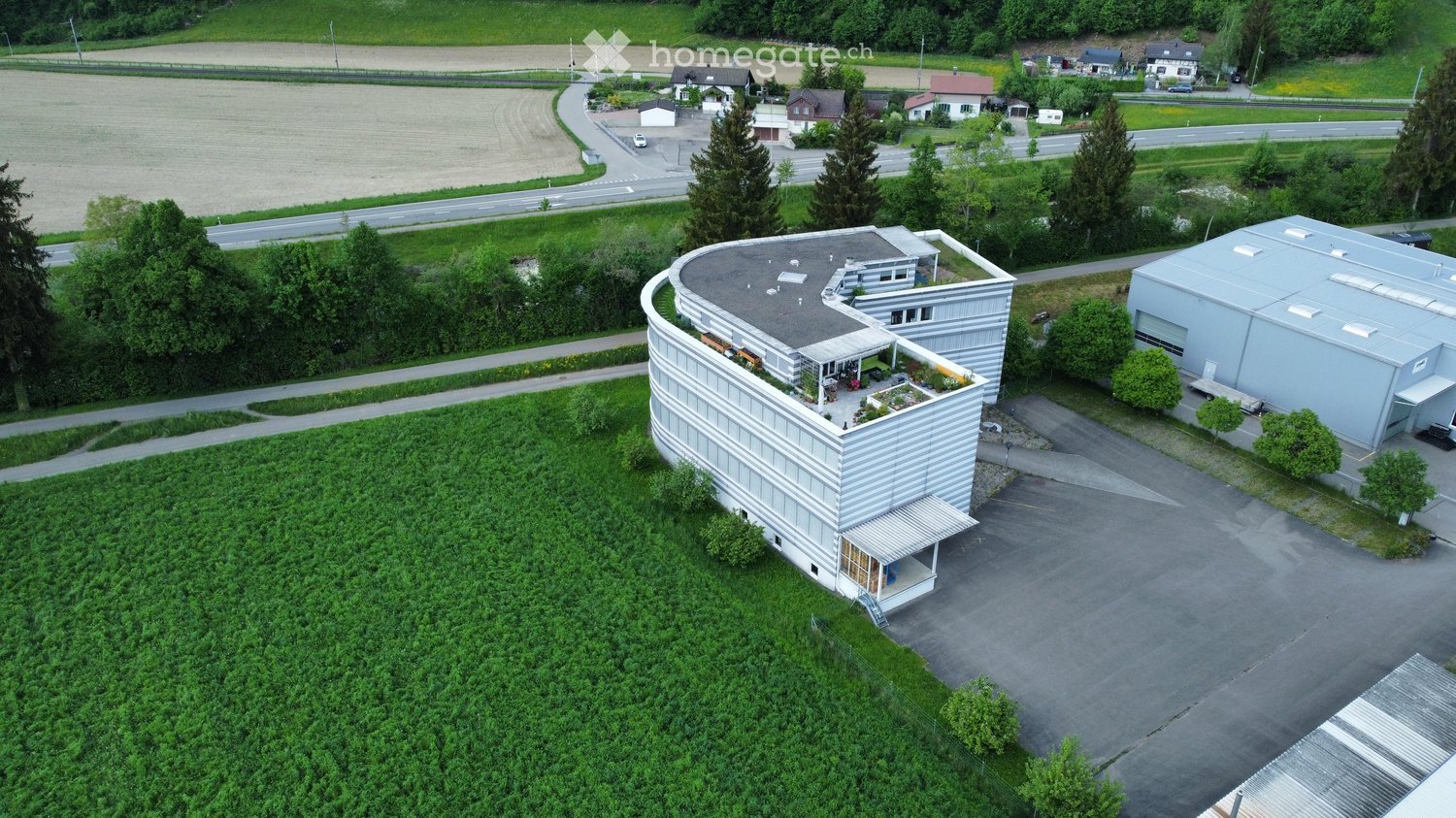 Building. A modern, curved building with a flat roof and a small garden area on the roof, surrounded by greenery and a parking lot.