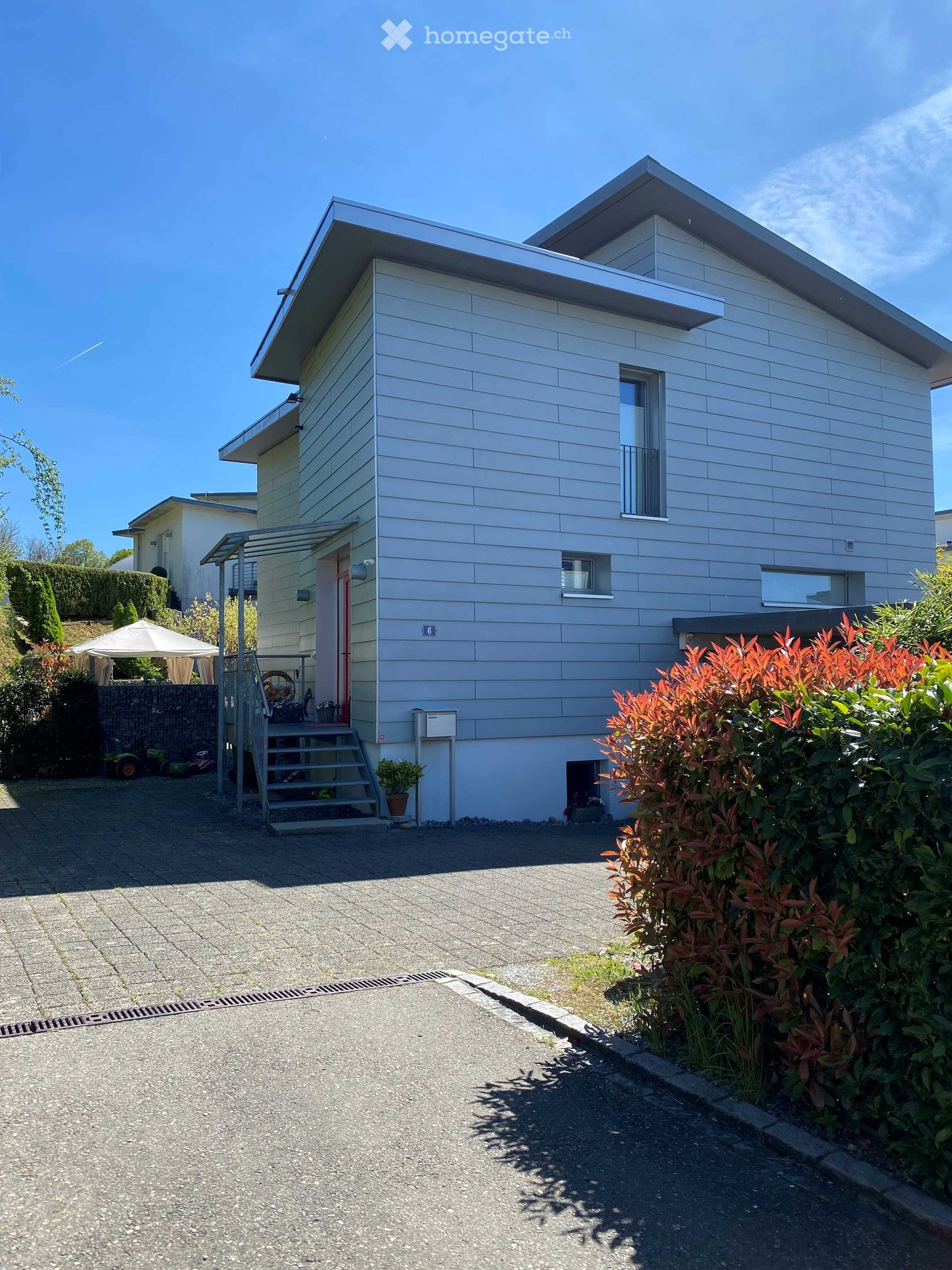House. Two-story house with light grey siding, multiple windows, and a small balcony on the upper level. The house has a covered entrance and is surrounded by greenery and a paved driveway.