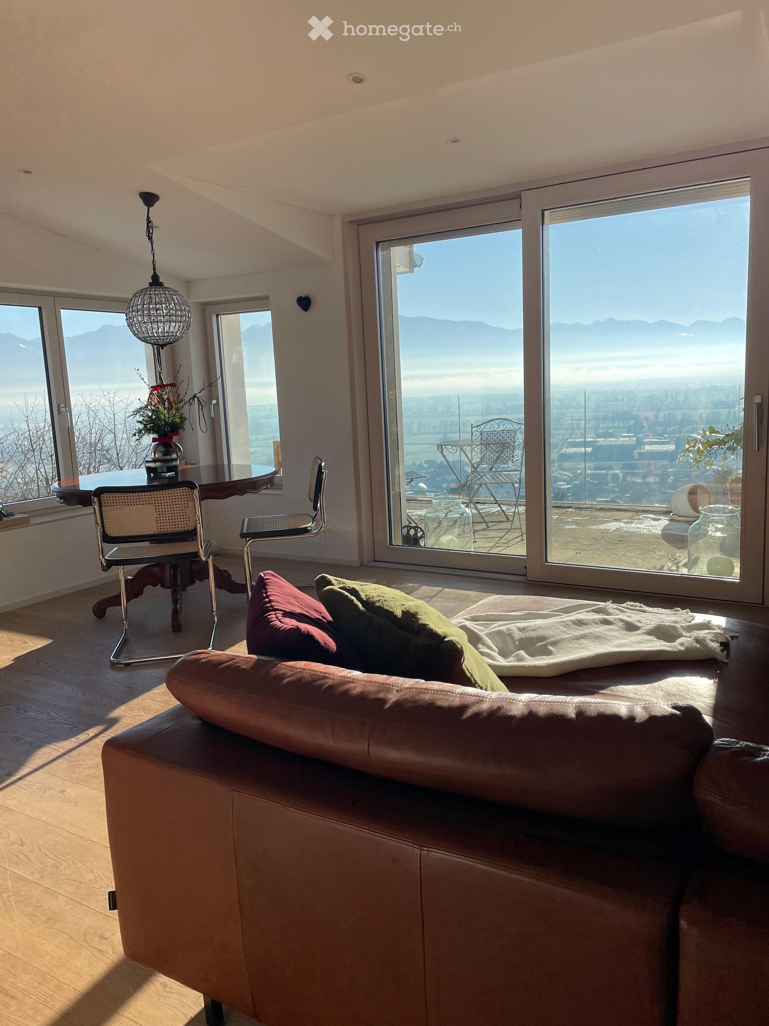 Living Room. Modern living room with large windows offering a scenic view, featuring a brown leather sofa, wooden floor, and a round table with chairs.