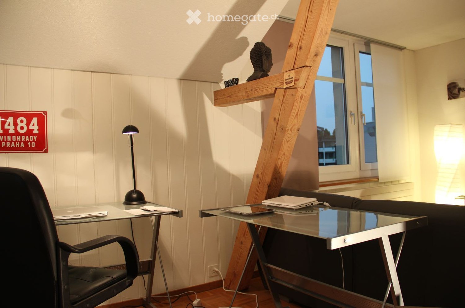 Furnished Room. A modern, well-lit room with a wooden slanted ceiling, featuring a black leather chair, a glass desk with a laptop, a small table with a lamp, and a sofa.