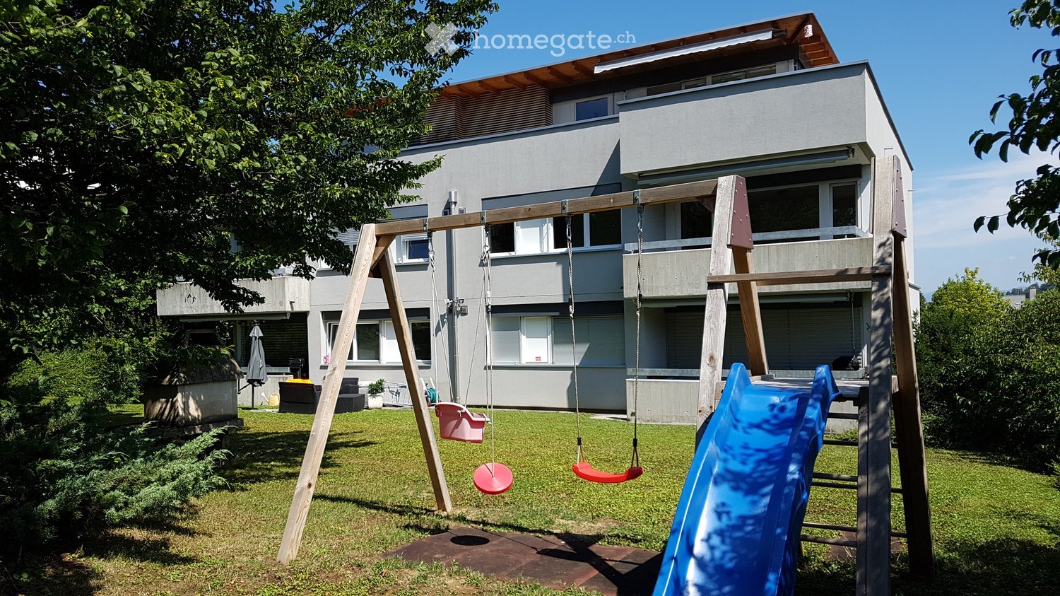 Two story house, concrete and wood playground equipment with a slide, swings, and bench.