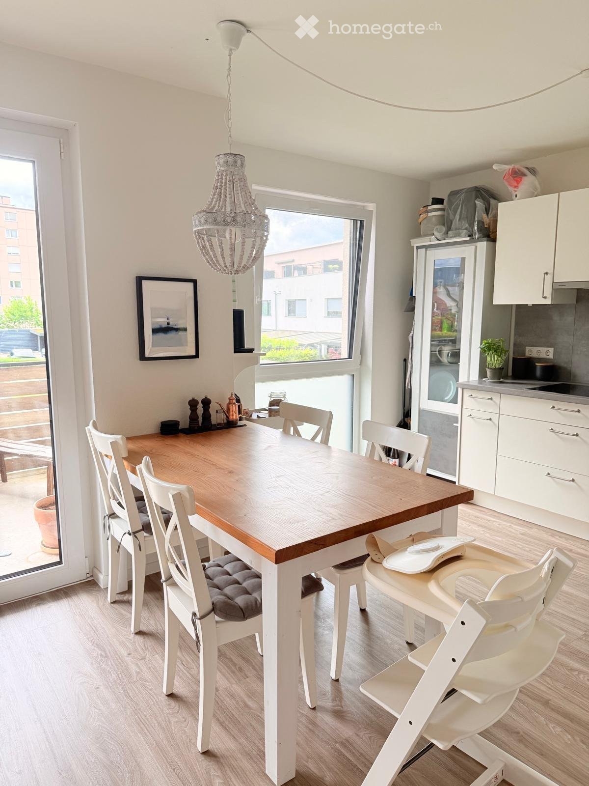 Kitchen. Bright kitchen and dining area with a wooden dining table, white chairs, a baby chair, modern kitchen cabinets, and a chandelier hanging from the ceiling. Large windows provide natural light and a view of the outside.