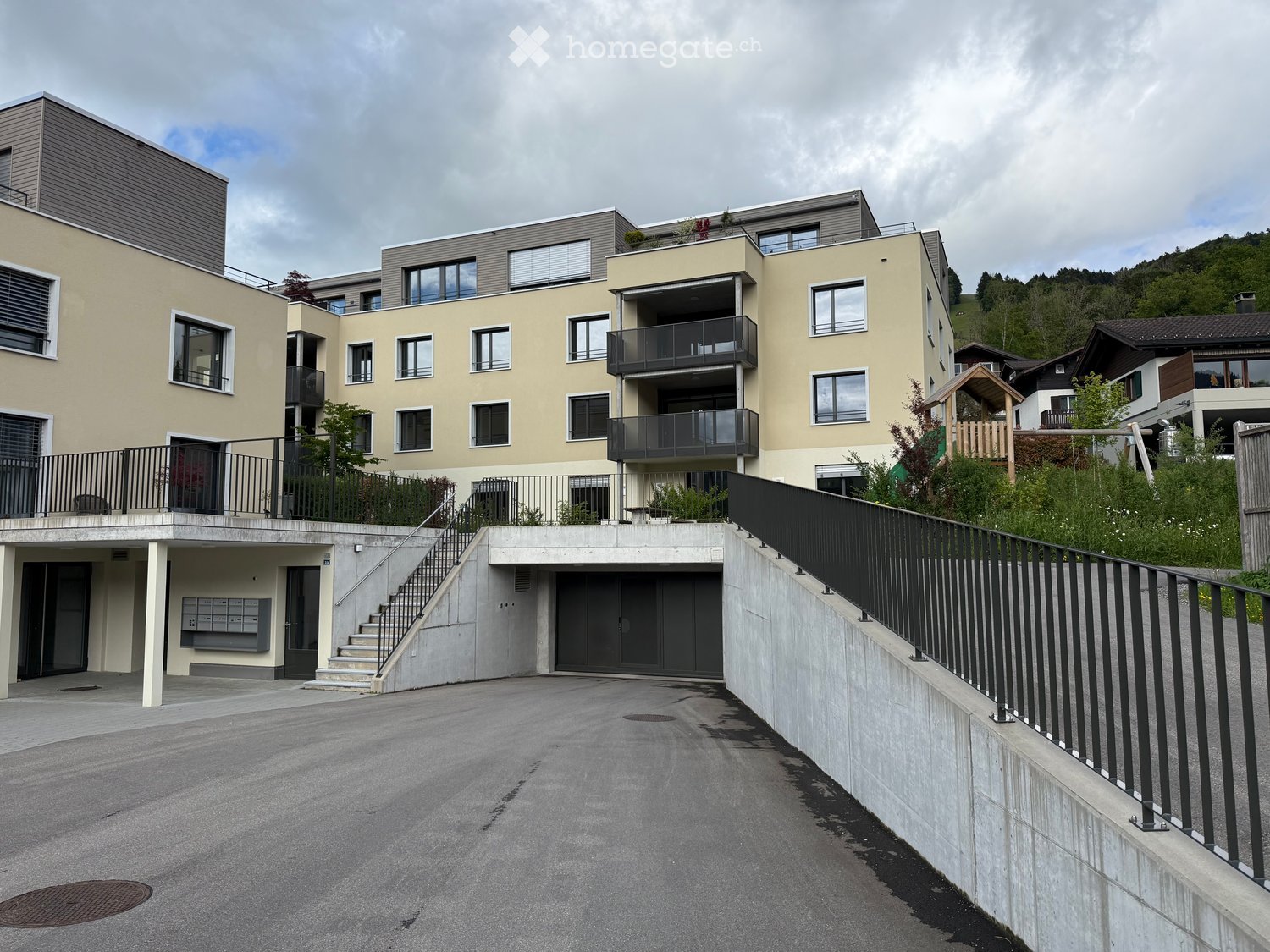 Building. Modern multi-story residential building with beige facade, large windows, balconies, and a raised ground floor with parking area.
