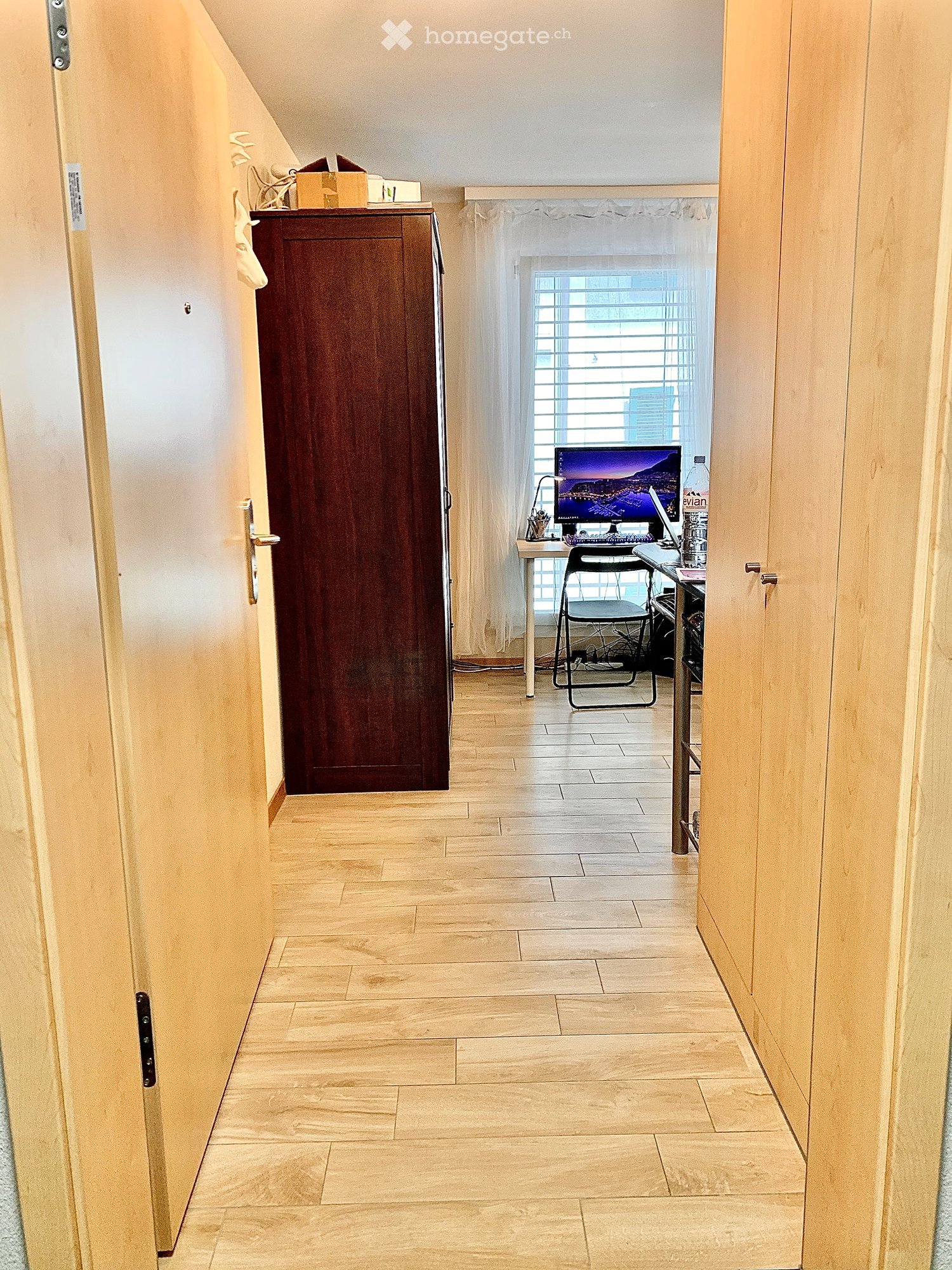 Corridor / Hallway. A narrow hallway with wooden flooring and light-colored walls, leading to a room with a desk, chair, and computer setup.