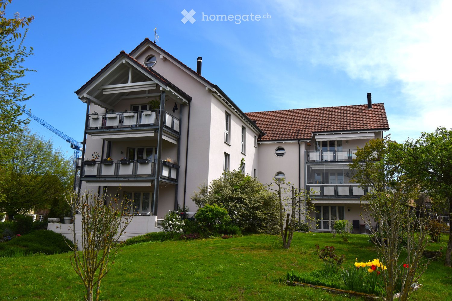 House. Two-story house with white exterior, multiple balconies, and a well-maintained garden with green lawn and colorful flowers.