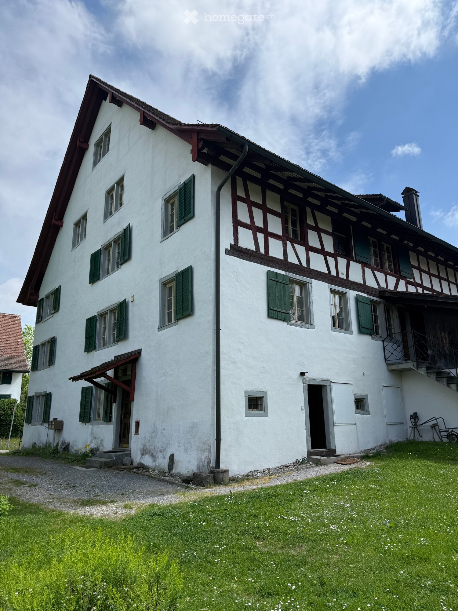 House. Two-story white house with green shutters, wooden balcony, and gravel driveway.