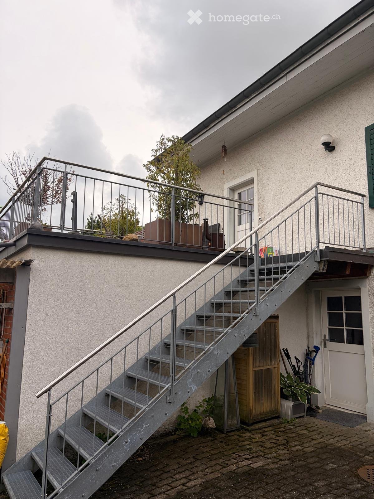 Building. Exterior view of a two-story house with a metal staircase leading to a rooftop terrace. The house has a light-colored facade, a small balcony, and some potted plants visible on the terrace.