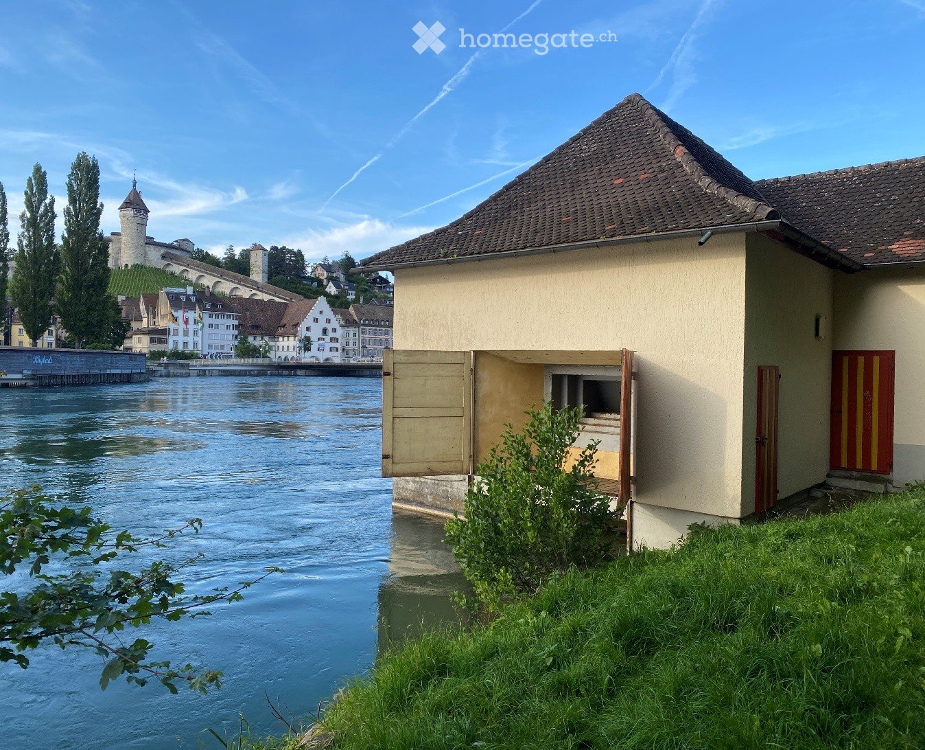 Building. A small beige building with a tiled roof sits near a river, with a view of a town and a castle in the background.