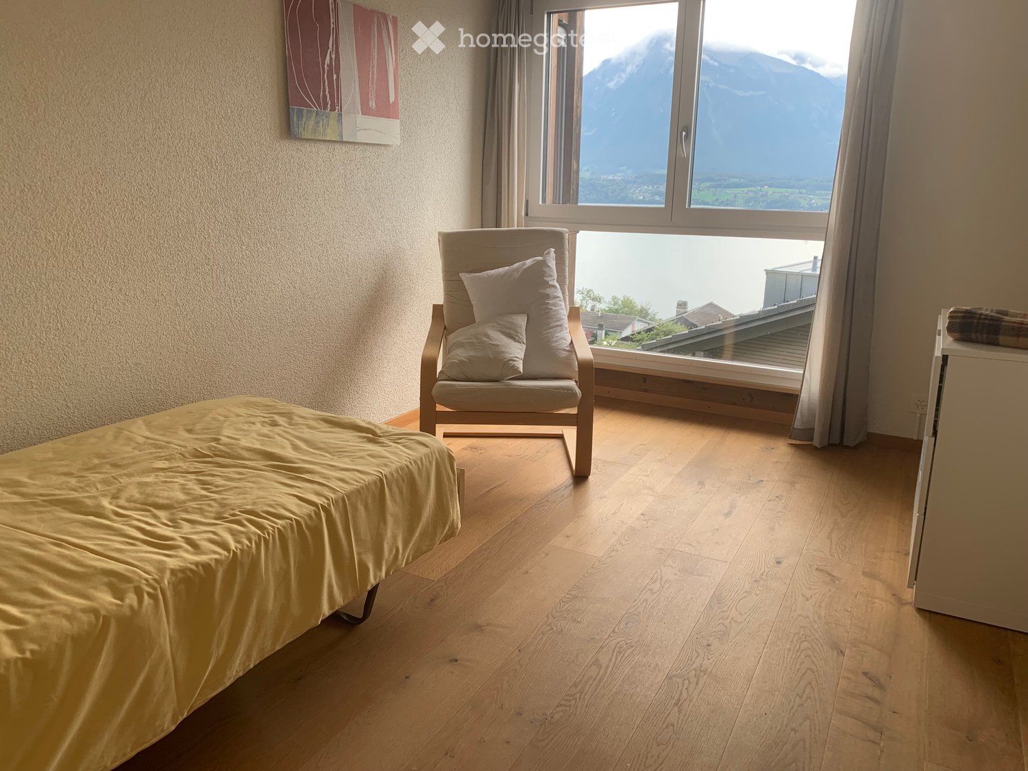 Bedroom. Bright bedroom with wooden flooring, a neatly made bed, a wooden armchair with pillows, and large windows offering a scenic mountain view.